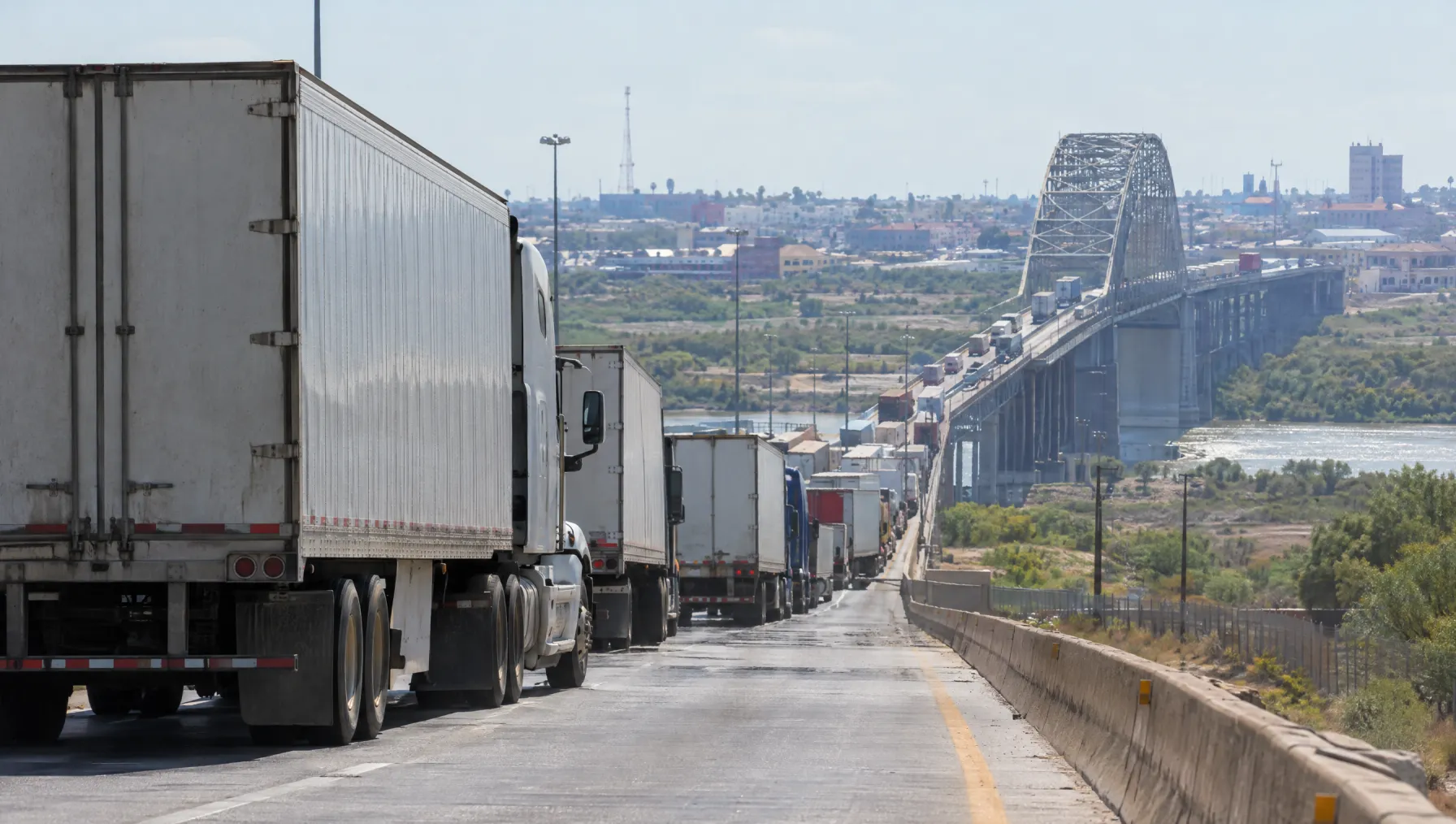 Class 8 tractor-trailers lined up in the commercial lanes approaching the World Trade Bridge in Laredo, Texas, with the Rio Grande and Nuevo Laredo in the distance under a hot afternoon sky